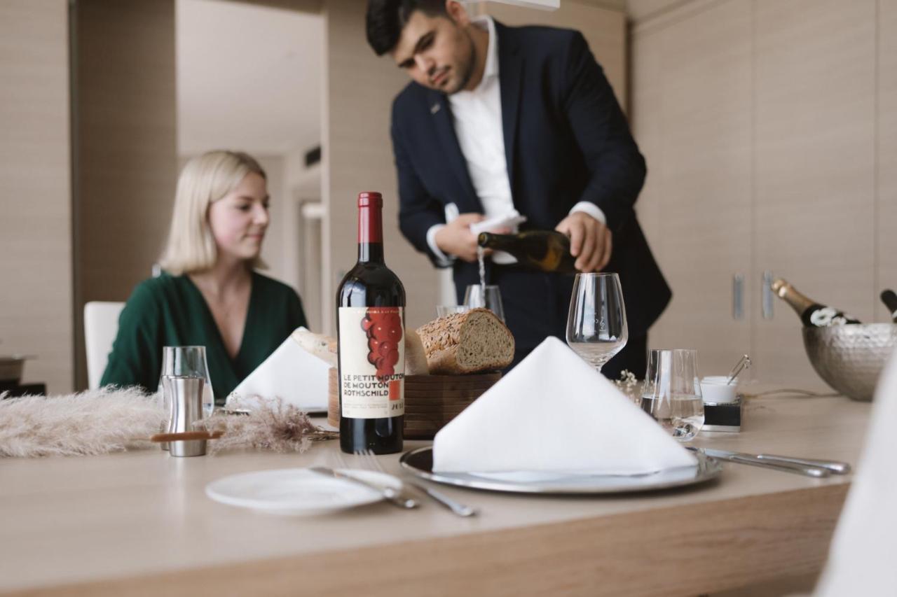 מלון The Westin Hamburg Elbphilharmonie מראה חיצוני תמונה The photo shows a dining scene featuring a well-dressed man pouring wine into a glass while a woman sits at the table. The table is elegantly set with a folded napkin, wine glasses, and a bottle of red wine placed prominently. There is also a loaf of