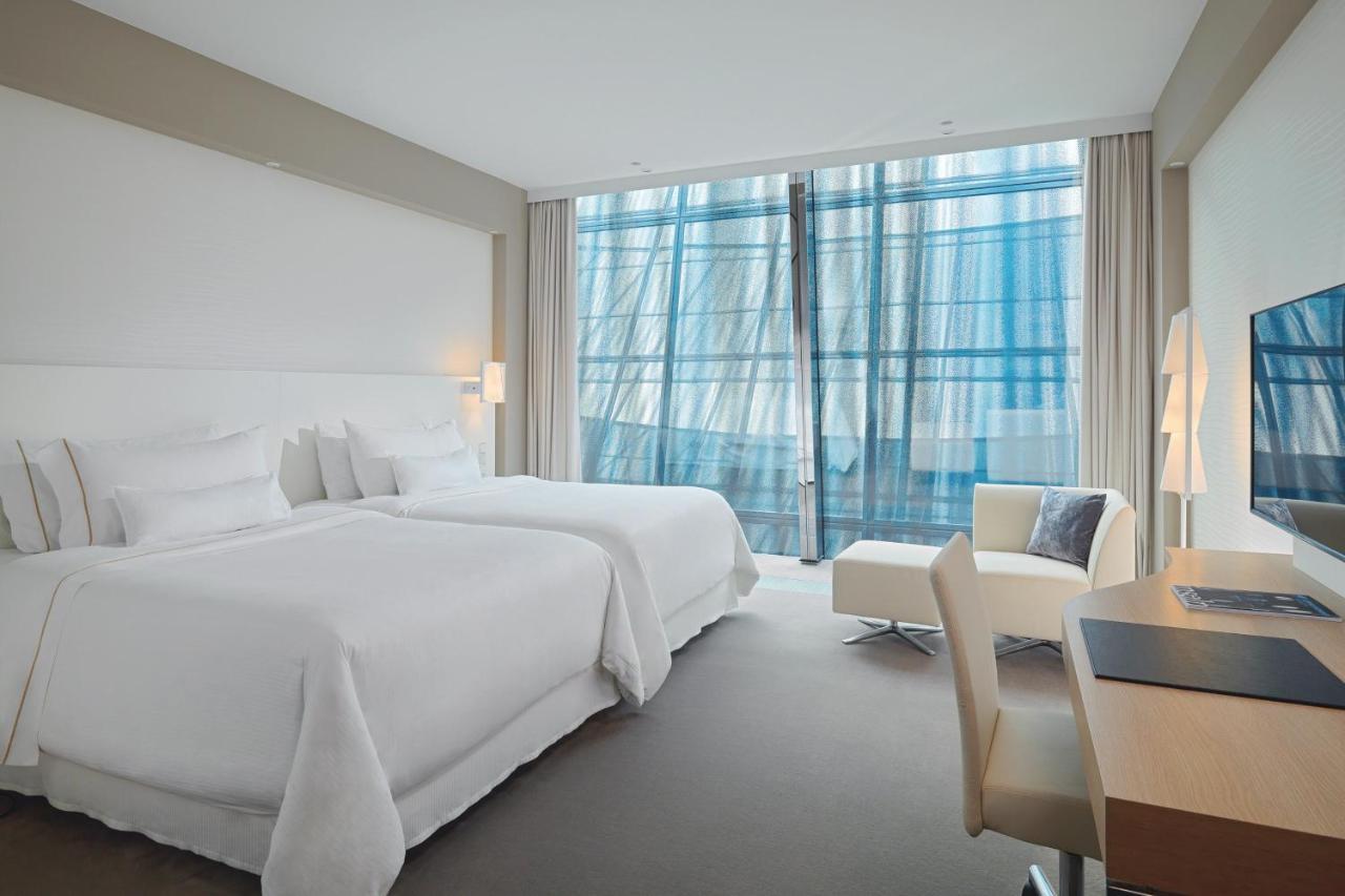 מלון The Westin Hamburg Elbphilharmonie מראה חיצוני תמונה The photo shows a modern hotel room featuring two white beds with plush bedding. There is a large window with sheer curtains that allows natural light to filter in, offering a view outside. The room has neutral-colored walls and carpeting, creating a