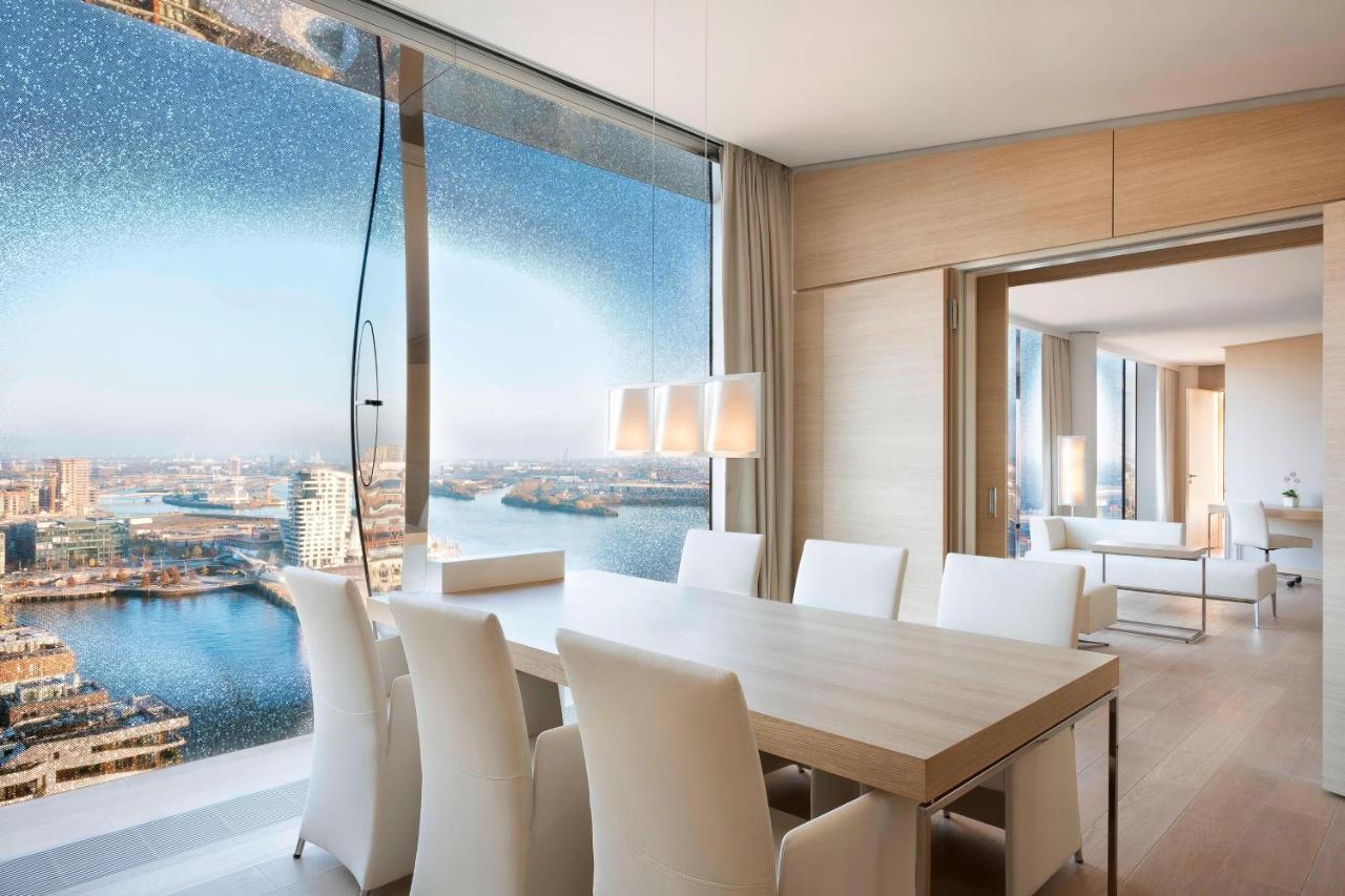 מלון The Westin Hamburg Elbphilharmonie מראה חיצוני תמונה The photo shows a modern dining area with a large, light-colored table surrounded by white chairs. The space features a panoramic window that offers a stunning view of a body of water and a cityscape. The decor is minimalistic and elegant, with smoot