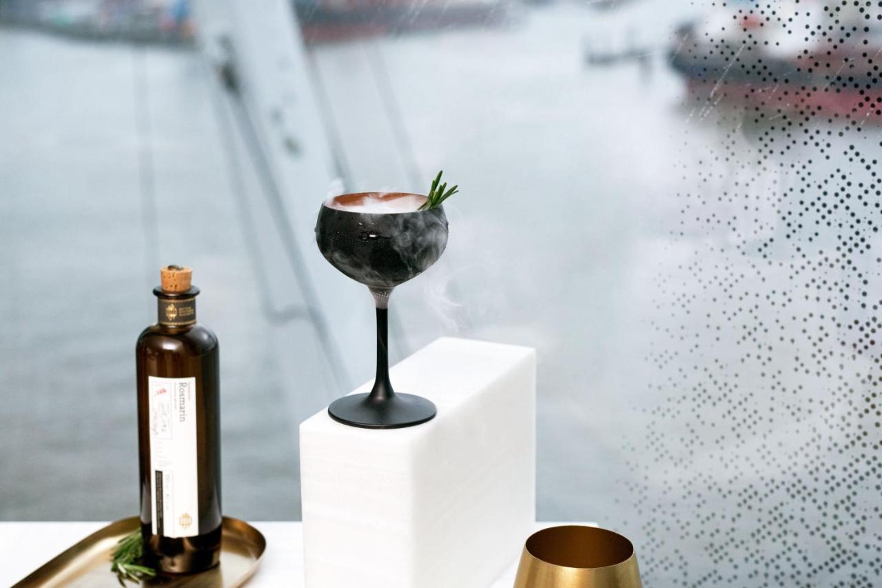מלון The Westin Hamburg Elbphilharmonie מראה חיצוני תמונה The photo features a stylish cocktail elegantly presented on a white pedestal. The cocktail is served in a sophisticated black stem glass, topped with a garnish of rosemary. Beside it, there is a dark glass bottle, likely containing a spirit or mixer