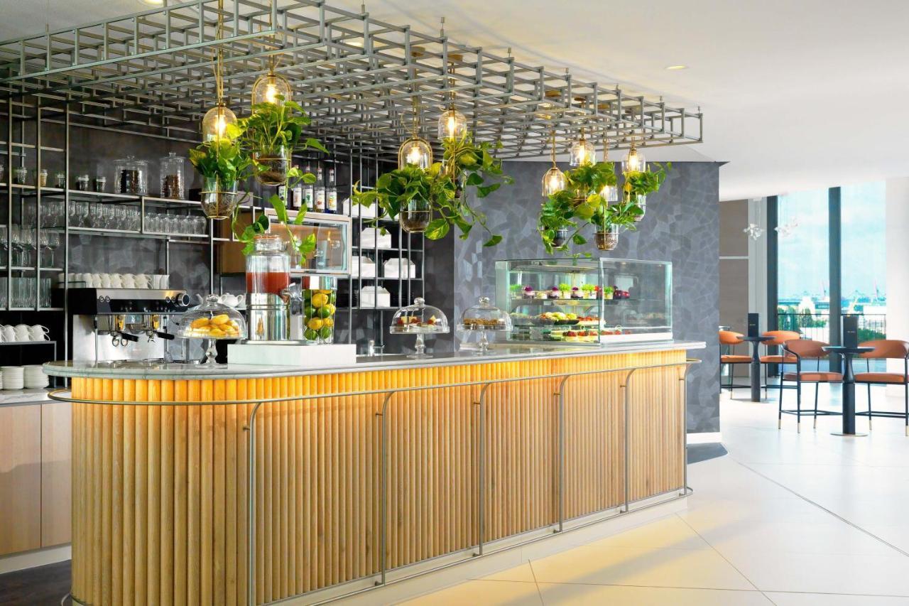 מלון The Westin Hamburg Elbphilharmonie מראה חיצוני תמונה The photo shows a modern café or bar setting with a sleek wooden counter that features vertical slats. Above the counter, there is a grid-like framework hanging from the ceiling, adorned with various green plants that add a touch of nature to the int