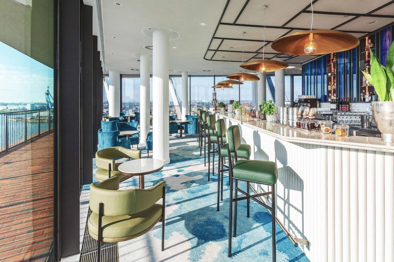 מלון The Westin Hamburg Elbphilharmonie מראה חיצוני תמונה The photo depicts a modern bar or lounge area with a stylish design. There are high-backed green chairs surrounding small circular tables, arranged in an open space that has large windows showcasing a view of the outside. The bar area is sleek, featu