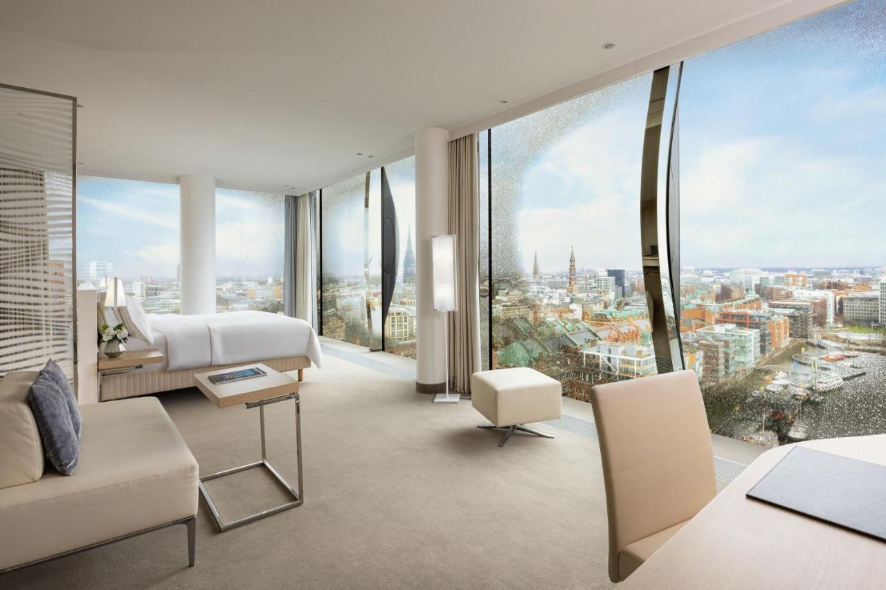 מלון The Westin Hamburg Elbphilharmonie מראה חיצוני תמונה The photo shows a modern hotel room with large floor-to-ceiling windows that provide a panoramic view of a city landscape. The room features a minimalist design with neutral tones. There's a king-sized bed positioned in the center, a sleek desk and c