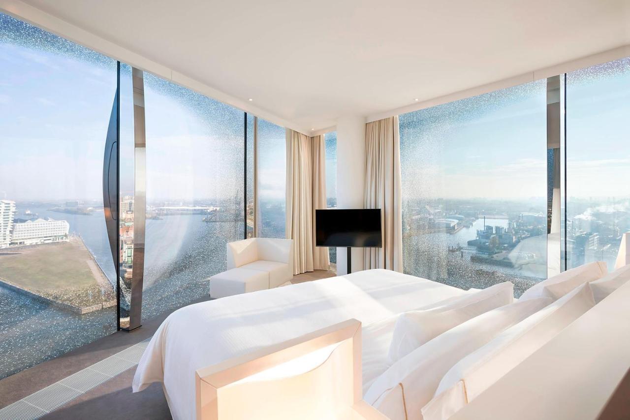 מלון The Westin Hamburg Elbphilharmonie מראה חיצוני תמונה The photo shows a modern hotel room featuring a large bed with white linens and a minimalist design. It has expansive floor-to-ceiling windows that offer a panoramic view of the landscape outside, likely a city or waterfront setting. The room is deco