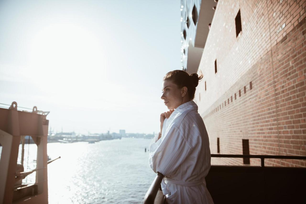 מלון The Westin Hamburg Elbphilharmonie מראה חיצוני תמונה The photo depicts a person standing on a balcony, looking out over a body of water. The individual is wearing a white bathrobe, and their hair is styled in a bun. The background features a scenic view with sunlight illuminating the scene, creating a 