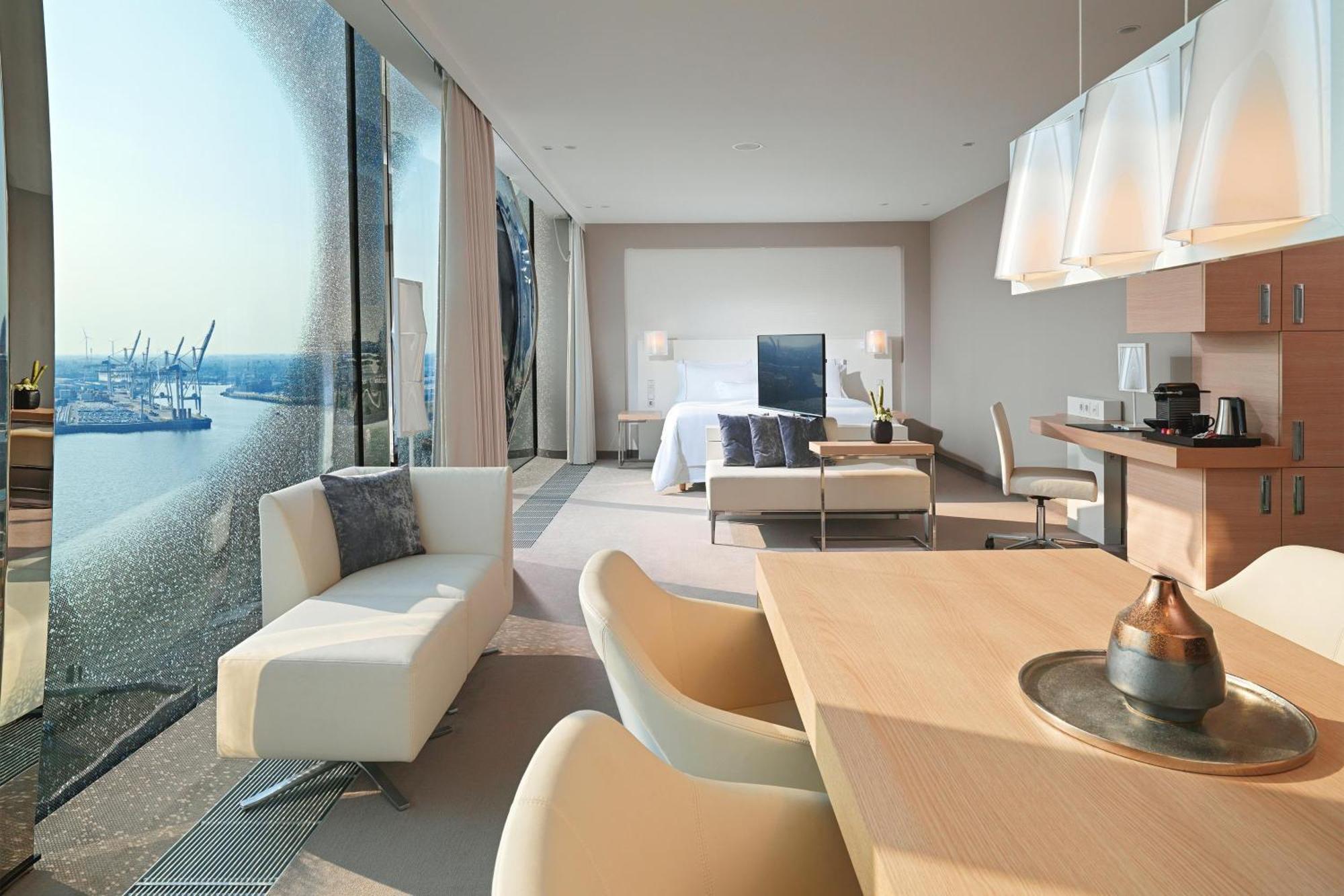 מלון The Westin Hamburg Elbphilharmonie מראה חיצוני תמונה The photo shows a modern hotel room with a spacious layout. On one side, there are large floor-to-ceiling windows that offer a view of the outside, likely showcasing a waterfront or cityscape. Inside, there's a bed with a minimalist design, accompani