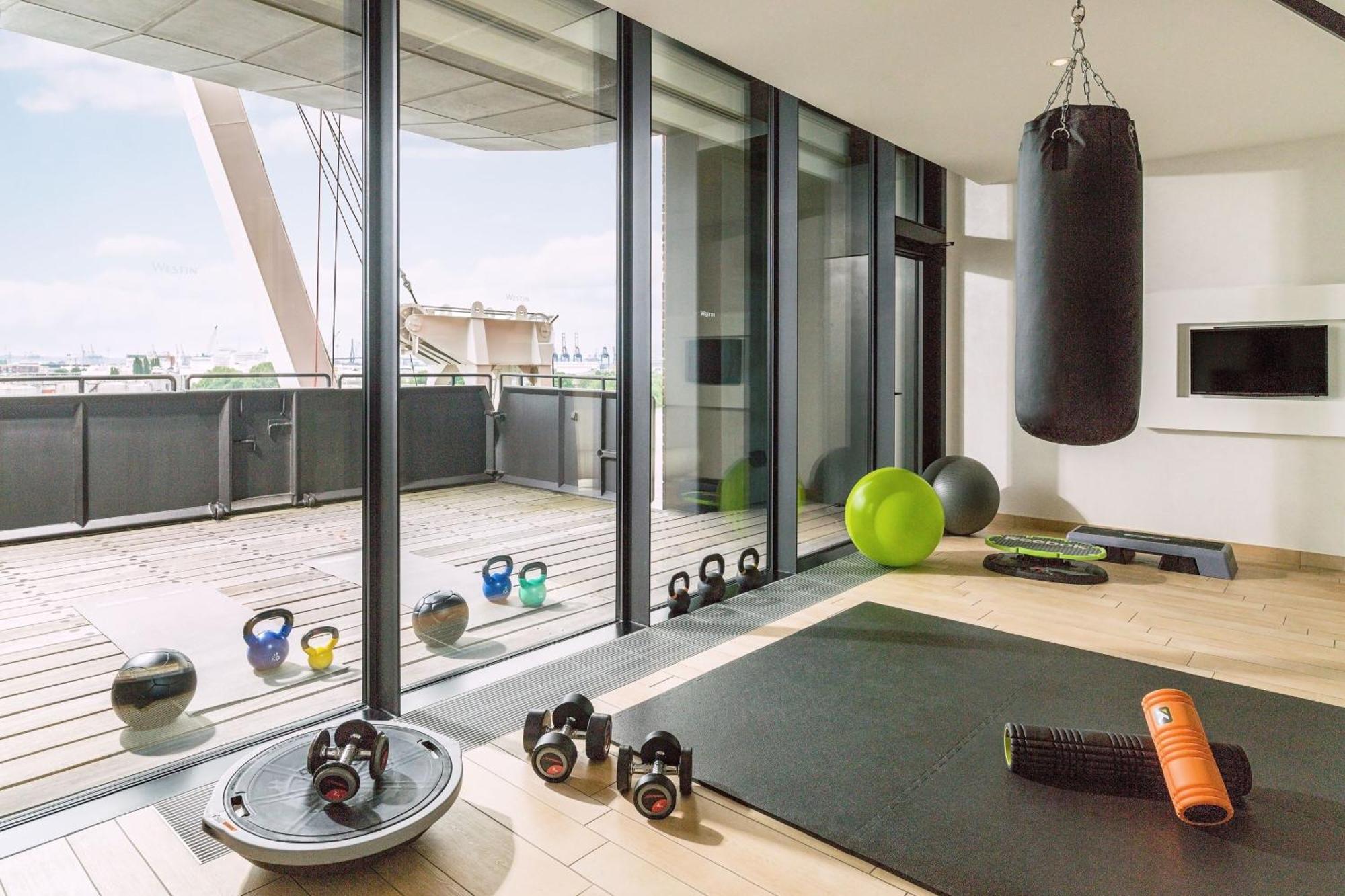 מלון The Westin Hamburg Elbphilharmonie מראה חיצוני תמונה The photo shows a modern home gym with large glass windows that provide a view to the outside. Inside, there are various fitness equipment items, including a heavy punching bag hanging from the ceiling, kettlebells, a stability ball, an exercise mat,