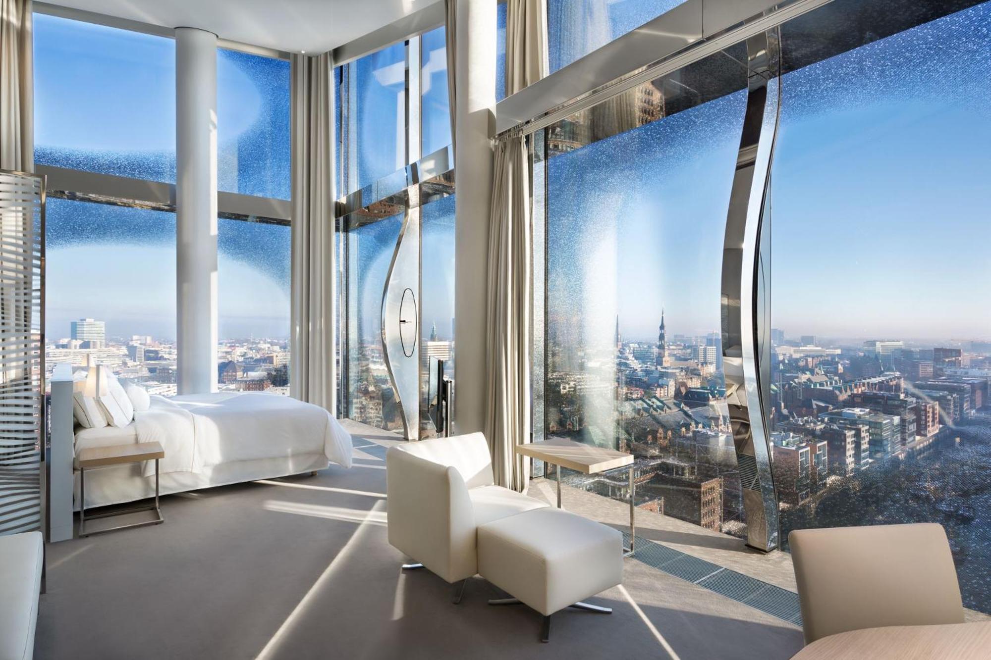 מלון The Westin Hamburg Elbphilharmonie מראה חיצוני תמונה The photo shows a modern hotel room with a stunning, panoramic view through large, uniquely designed windows. The room features a sleek, minimalist aesthetic with light-colored furnishings, including a white bed and a couple of chairs. The walls are 